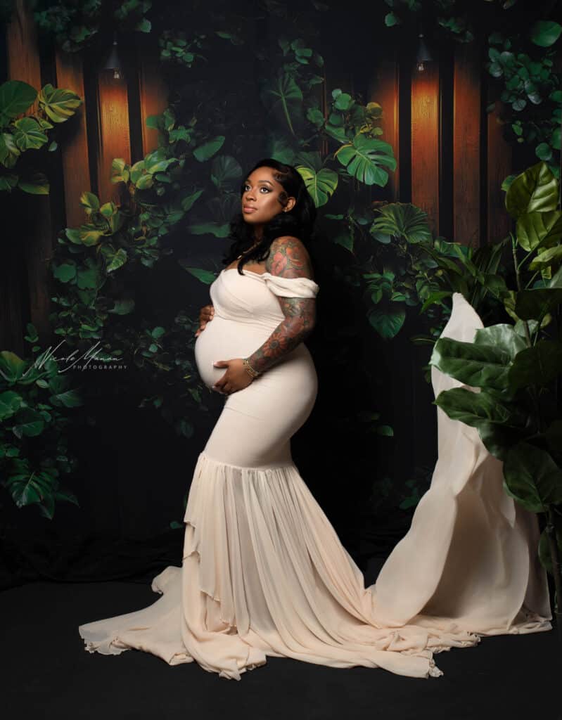 Woman during her maternity session in Columbus GA in a white dress with a long tossing train, she is standing in front of lots of tropical plants.