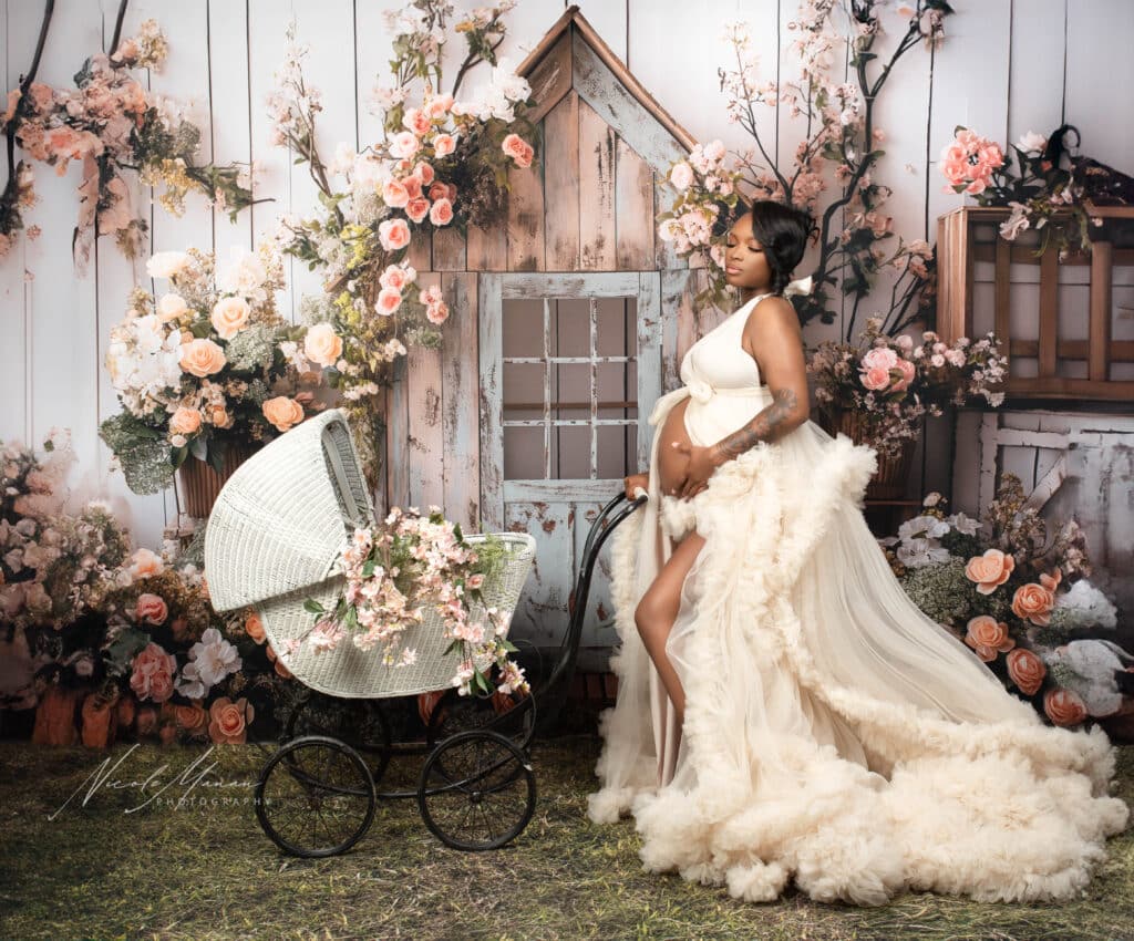 Woman during her maternity session in Columbus GA in a white dress with lots of ruffles, the atmosphere is romantic with lots of flowers and a little garden shed. She has her hands resting on white vintage baby carriage.