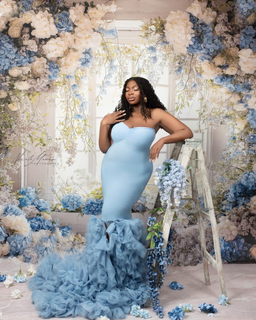 Woman wearing a blue gown during her maternity session in Columbus GA leaning against a white ladder. She is standing in front of a large window decorated in lots of blue and white flowers.