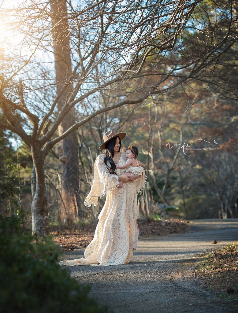 A mother holding her child in a loving embrace in an fall photo shoot, she is wearing a dreamy white lace boho gown and a brown head and smiling at her baby.