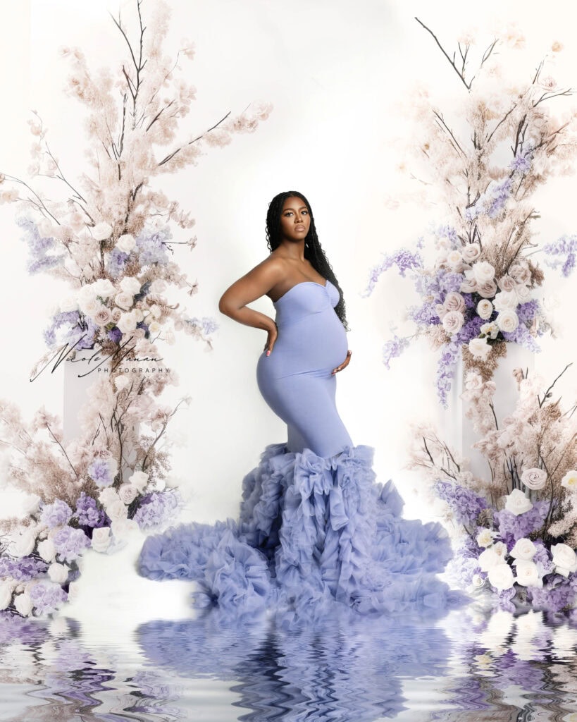 Pregnant black woman in a beautiful posed during her maternity photo shoot in Columbus GA. She is wearing a lavender color gown with ruffles that is very elegant and standing in the middle of lavender and while floral decoration.