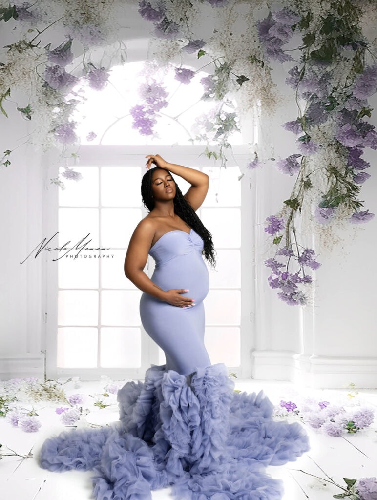Elegant maternity portrait of a pregnant woman in a lavender gown with ruffled train, floral decor, and soft natural lighting.