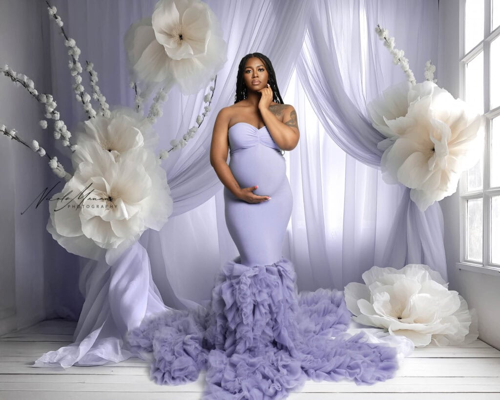 Elegant pregnant woman in lavender gown with ruffled train, surrounded by large white floral decorations, in a softly lit studio.