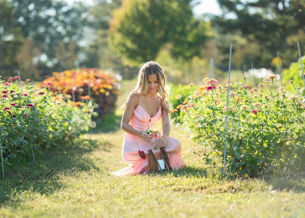 A girl in a long flowy pink dress sitting in a flower field during golden hour. She is cutting flowers and adding them to her wooden bucket, the sun is in her hair, during her golden hour senior session in Columbus, Ga.