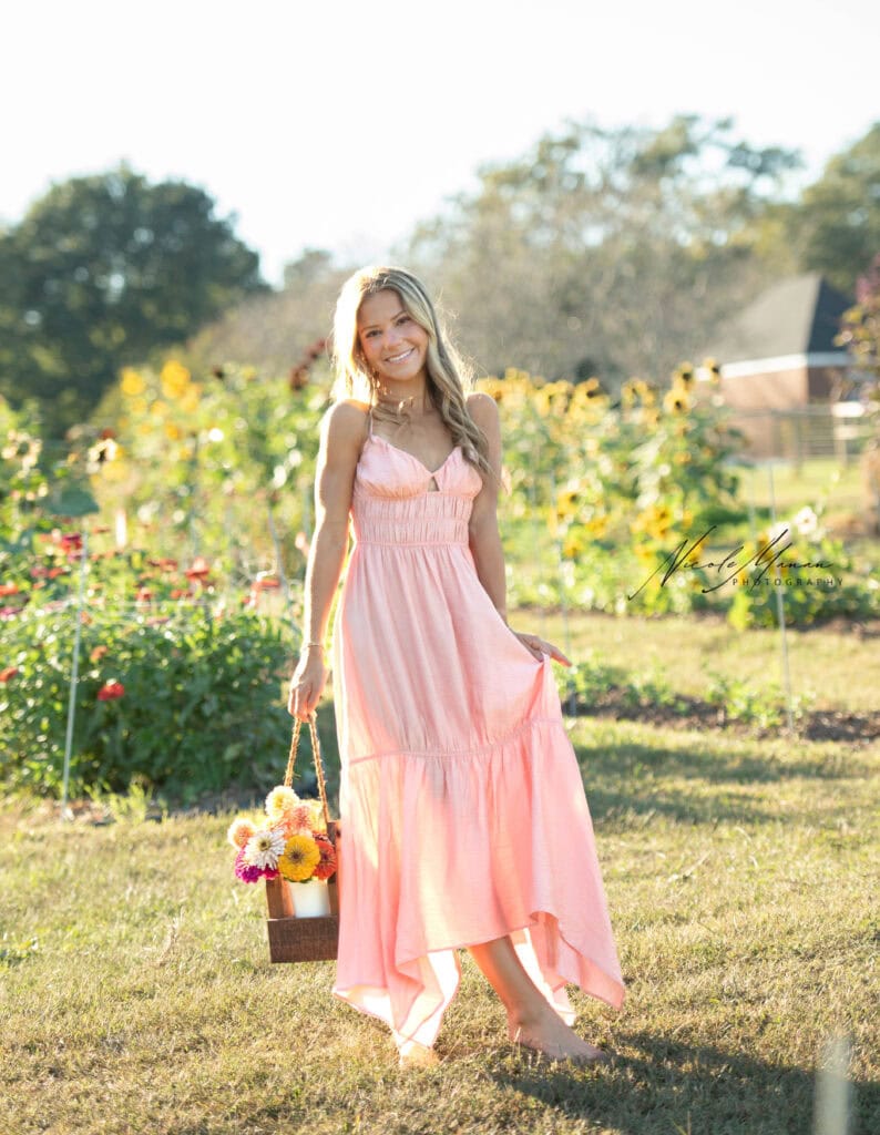 A girl in a long flowy pink dress standing in a flower field during golden hour. She is holding a bucket filled with colorful flowers during her senior session in Columbus, Ga.