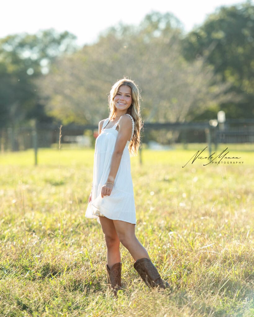 A girl in a white dress and cowboy boots in a wheat field at golden hour during her senior session., smiling at the camera with the golden light in her hair.