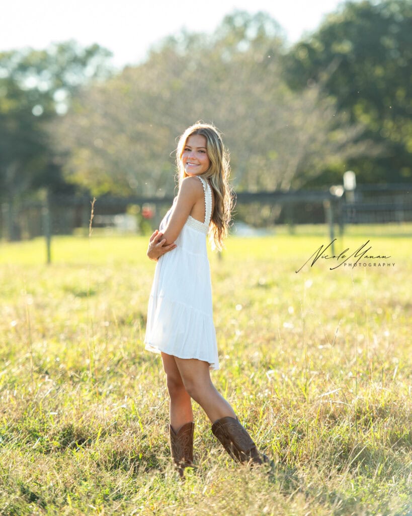 A girl in a white dress and cowboy boots in a wheat field at golden hour during her senior session.