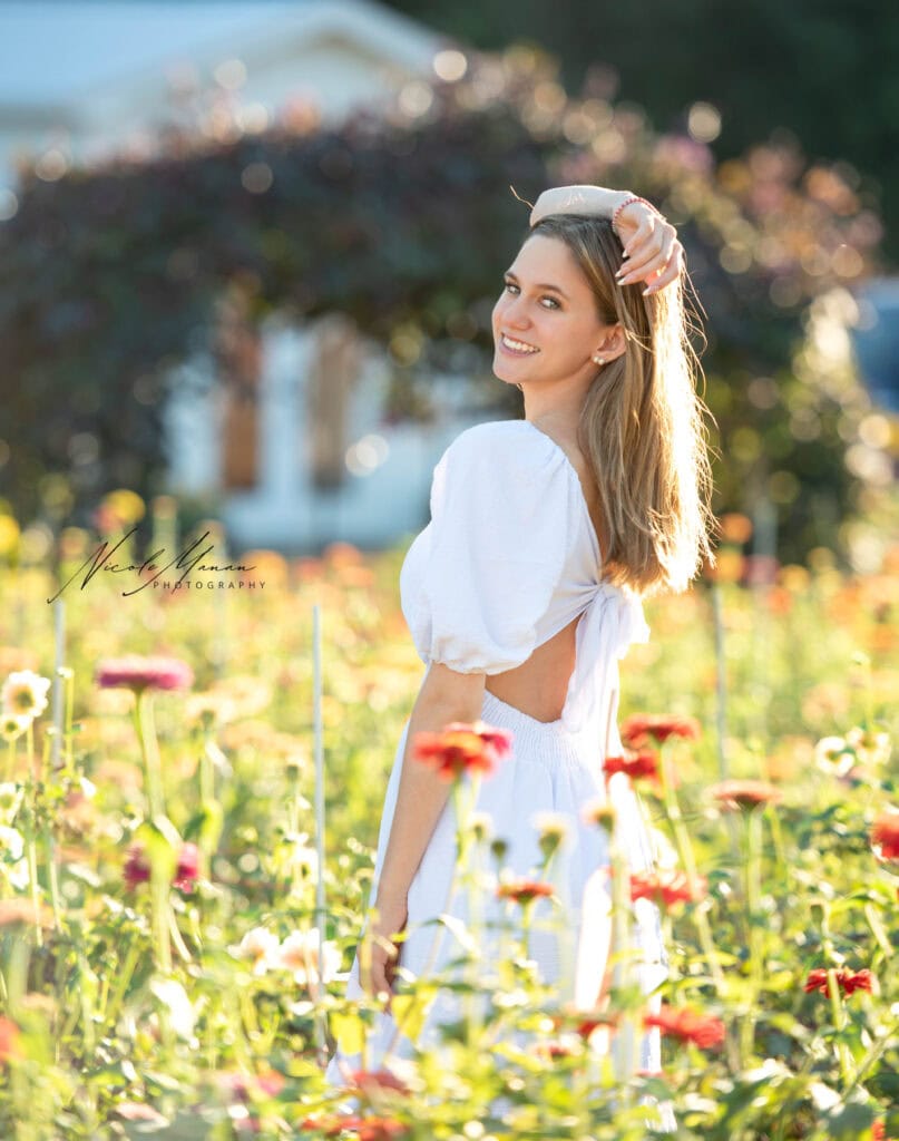A girl standing in a flower field with her hand over her head and smiling during her senior photo session.