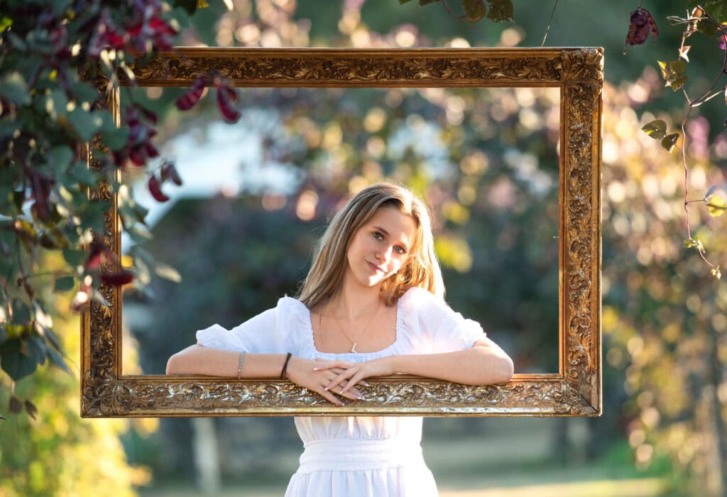 A girl resting her arms on a large golden frame hanging from a floral arch during her senior session in Columbus Ga.