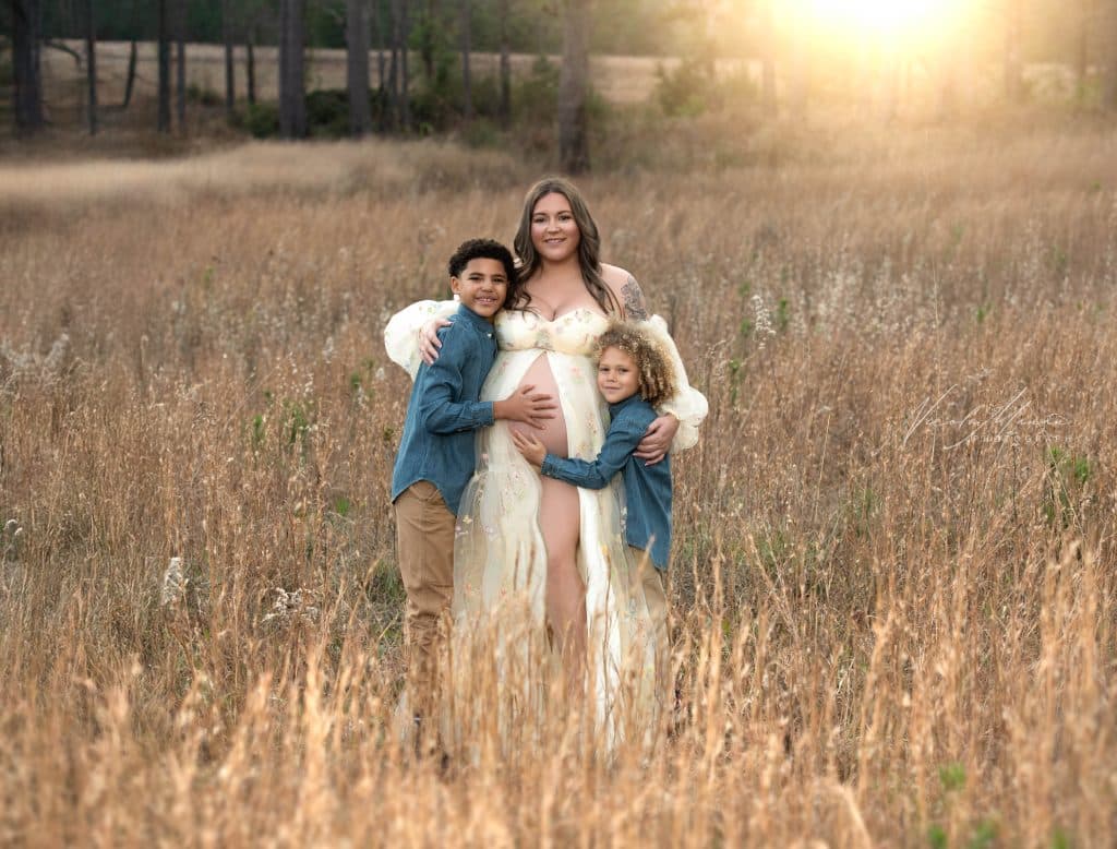 An pregnant woman in a white flowy dress with her 2 young children hugging her. She is standing in a wheat field for her maternity session at golden hour.